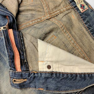 May include: Close-up of a denim jacket with a partially unzipped front. The zipper has a coral-colored stripe. The jacket shows signs of wear and tear, with visible stitching and a faded appearance. The interior lining is a light beige color.