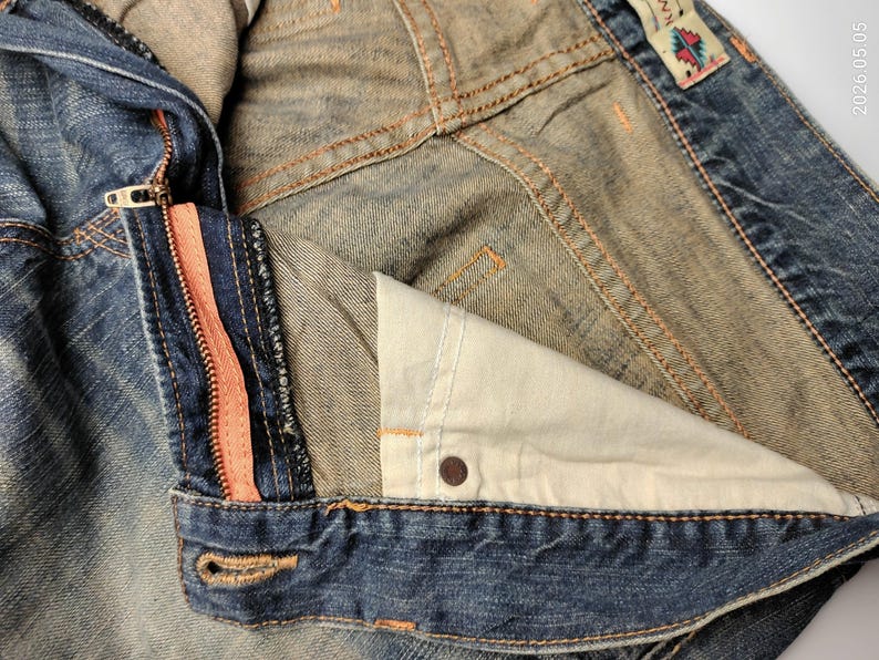 May include: Close-up of a denim jacket with a partially unzipped front. The zipper has a coral-colored stripe. The jacket shows signs of wear and tear, with visible stitching and a faded appearance. The interior lining is a light beige color.