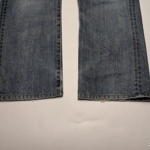 May include: Close-up of the bottom of a pair of blue denim jeans. The jeans have a classic five-pocket design with a straight leg and a medium wash. The hems are finished with a standard double-needle stitch.