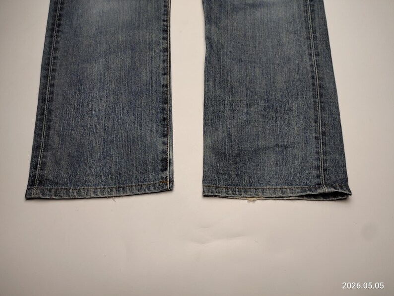 May include: Close-up of the bottom of a pair of blue denim jeans. The jeans have a classic five-pocket design with a straight leg and a medium wash. The hems are finished with a standard double-needle stitch.
