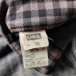 Puede incluir: Primer plano de una etiqueta de prenda vaquera con la marca "EDWIN" y la talla "28". La etiqueta est&aacute; adherida a una tela vaquera con forro de cuadros. La tela es de color azul descolorido con costuras naranjas.