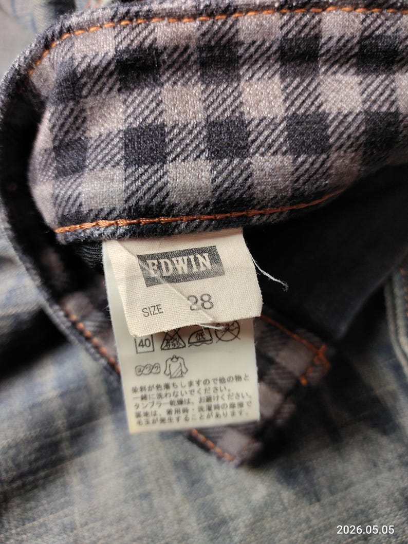 Puede incluir: Primer plano de una etiqueta de prenda vaquera con la marca "EDWIN" y la talla "28". La etiqueta est&aacute; adherida a una tela vaquera con forro de cuadros. La tela es de color azul descolorido con costuras naranjas.