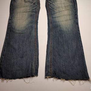 May include: A pair of blue denim jeans with a flared leg design. The jeans have a faded wash and distressed detailing, including frayed edges at the hem. The stitching is visible in a contrasting brown thread.