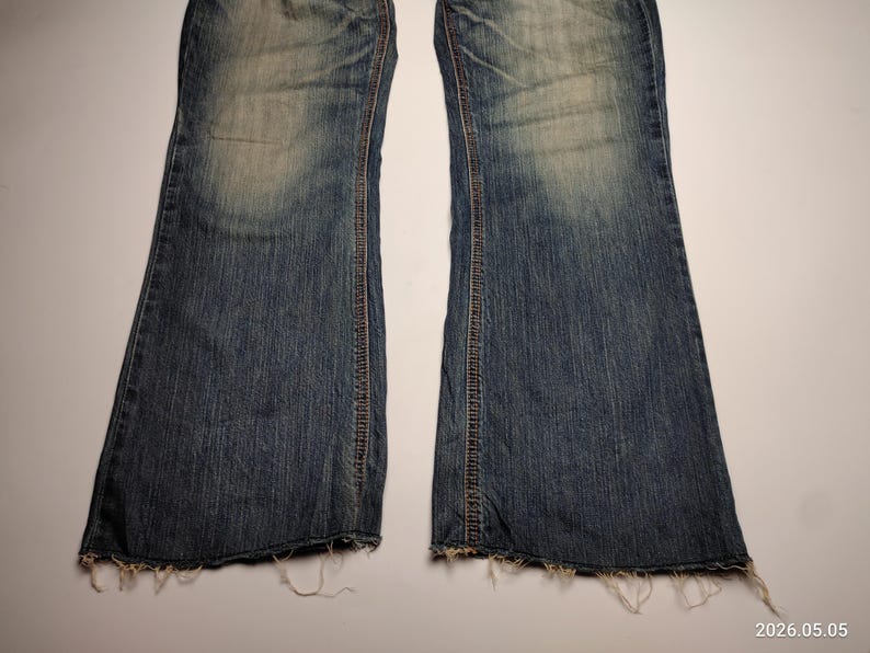 May include: A pair of blue denim jeans with a flared leg design. The jeans have a faded wash and distressed detailing, including frayed edges at the hem. The stitching is visible in a contrasting brown thread.