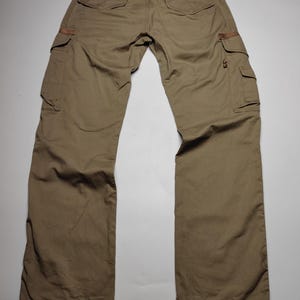 May include: Khaki-colored cargo pants with multiple pockets and brown leather accents. The pants have a relaxed fit and a classic design, suitable for casual wear. The back of the pants is visible, showing the pockets and belt loops.