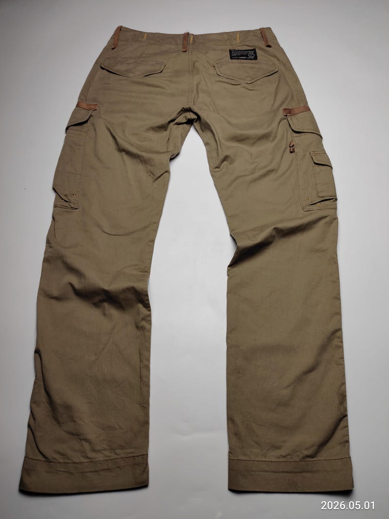 May include: Khaki-colored cargo pants with multiple pockets and brown leather accents. The pants have a relaxed fit and a classic design, suitable for casual wear. The back of the pants is visible, showing the pockets and belt loops.