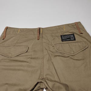 May include: Khaki-colored cargo pants with two back pockets and a small rectangular label. The pants have brown accents on the belt loops and side pockets. The fabric appears to be a durable, woven material.