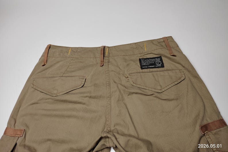 May include: Khaki-colored cargo pants with two back pockets and a small rectangular label. The pants have brown accents on the belt loops and side pockets. The fabric appears to be a durable, woven material.