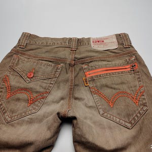 May include: Brown denim jeans with orange stitching and zipper pocket details. Features a classic five-pocket design with a branded leather patch. The jeans have a faded, worn look.