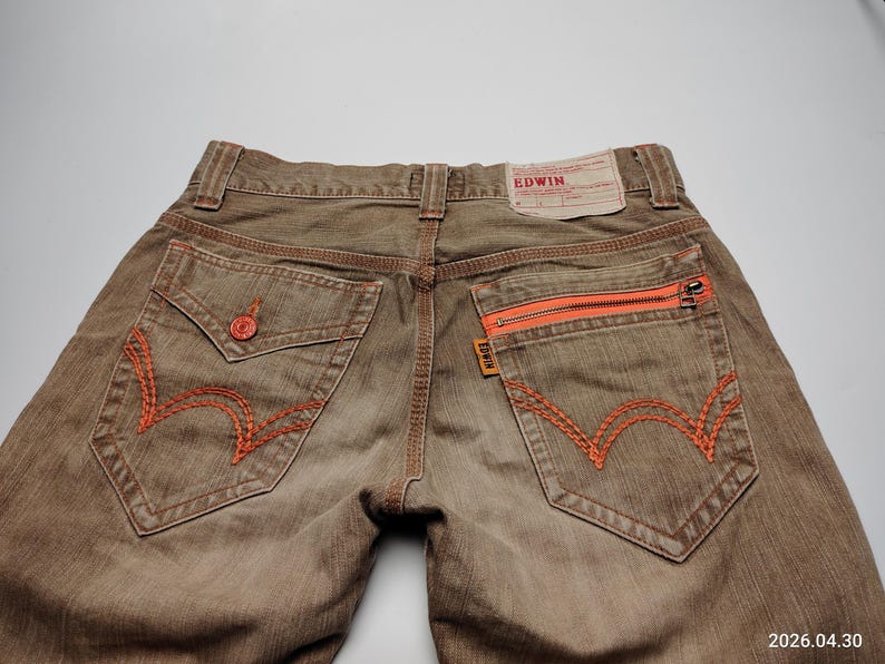 May include: Brown denim jeans with orange stitching and zipper pocket details. Features a classic five-pocket design with a branded leather patch. The jeans have a faded, worn look.