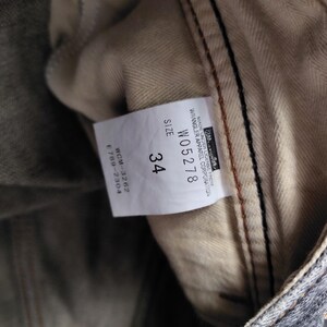May include: Close-up of a pair of denim jeans with a size 34 label. The label includes the Wrangler Apparel Corporation logo and product details. The jeans have a gray and beige color scheme.