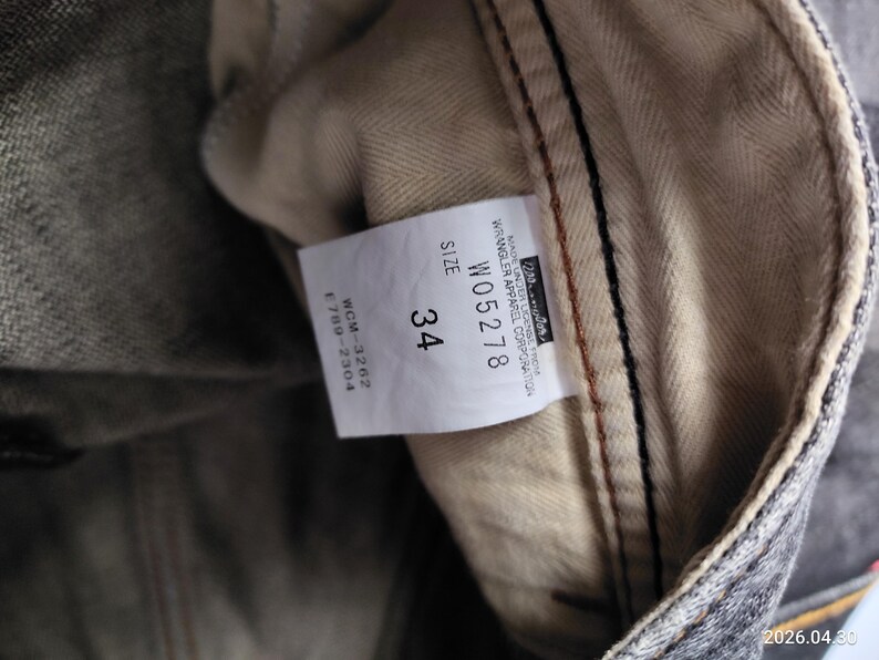 May include: Close-up of a pair of denim jeans with a size 34 label. The label includes the Wrangler Apparel Corporation logo and product details. The jeans have a gray and beige color scheme.