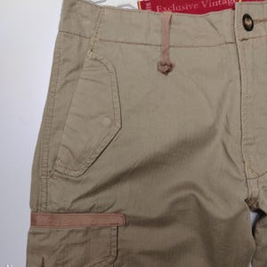 May include: Close-up of khaki-colored cargo shorts. The shorts feature multiple pockets, including a side cargo pocket with a brown accent. The waistband has a button closure and a small loop. The text "Exclusive Vintage" is visible.