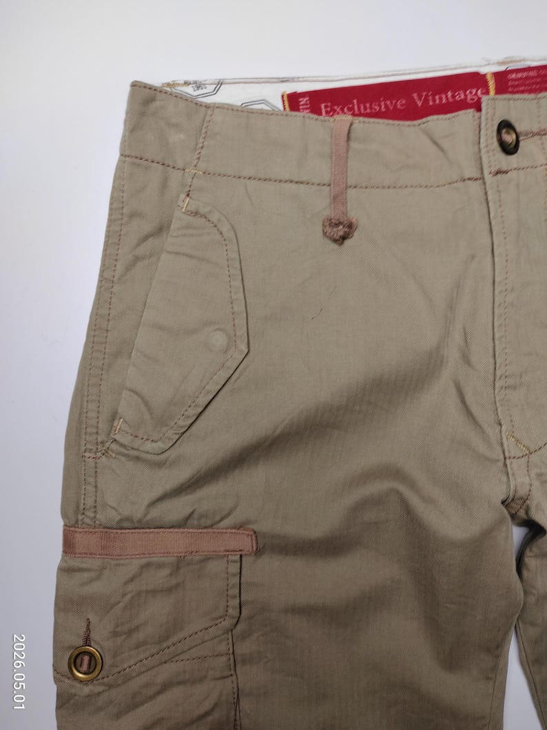 May include: Close-up of khaki-colored cargo shorts. The shorts feature multiple pockets, including a side cargo pocket with a brown accent. The waistband has a button closure and a small loop. The text "Exclusive Vintage" is visible.