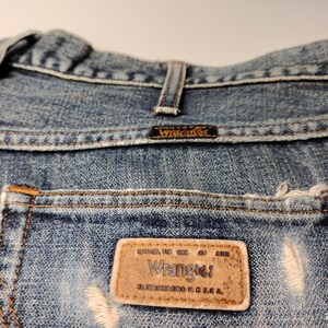Peut inclure: Gros plan sur un jean en denim bleu avec une poche us&eacute;e. L'&eacute;tiquette de la marque Wrangler est visible sur la poche arri&egrave;re, ainsi que le nom de la marque sur la ceinture. Le jean a un aspect classique et us&eacute;.