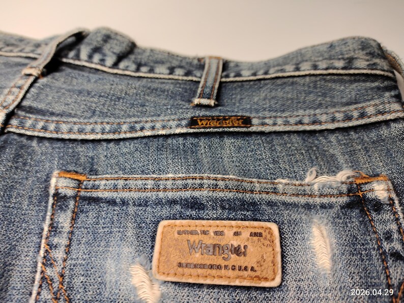 Peut inclure: Gros plan sur un jean en denim bleu avec une poche us&eacute;e. L'&eacute;tiquette de la marque Wrangler est visible sur la poche arri&egrave;re, ainsi que le nom de la marque sur la ceinture. Le jean a un aspect classique et us&eacute;.