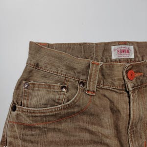 May include: Close-up of brown denim jeans with orange stitching and a button. The jeans have a classic five-pocket design and a brand label. The fabric appears to be slightly faded, giving it a vintage look. The jeans are a neutral color.