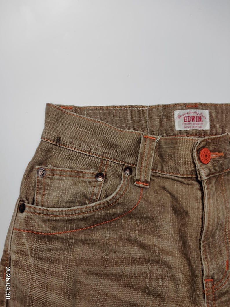 May include: Close-up of brown denim jeans with orange stitching and a button. The jeans have a classic five-pocket design and a brand label. The fabric appears to be slightly faded, giving it a vintage look. The jeans are a neutral color.