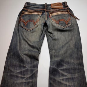 Puede incluir: Vaqueros de mezclilla de lavado oscuro con detalles en cuero marr&oacute;n y costuras. Incluyen bolsillos traseros decorativos con pespuntes naranjas, un parche de cuero marr&oacute;n y un detalle de cremallera. Los vaqueros tienen un aspecto descolorido y desgastado.