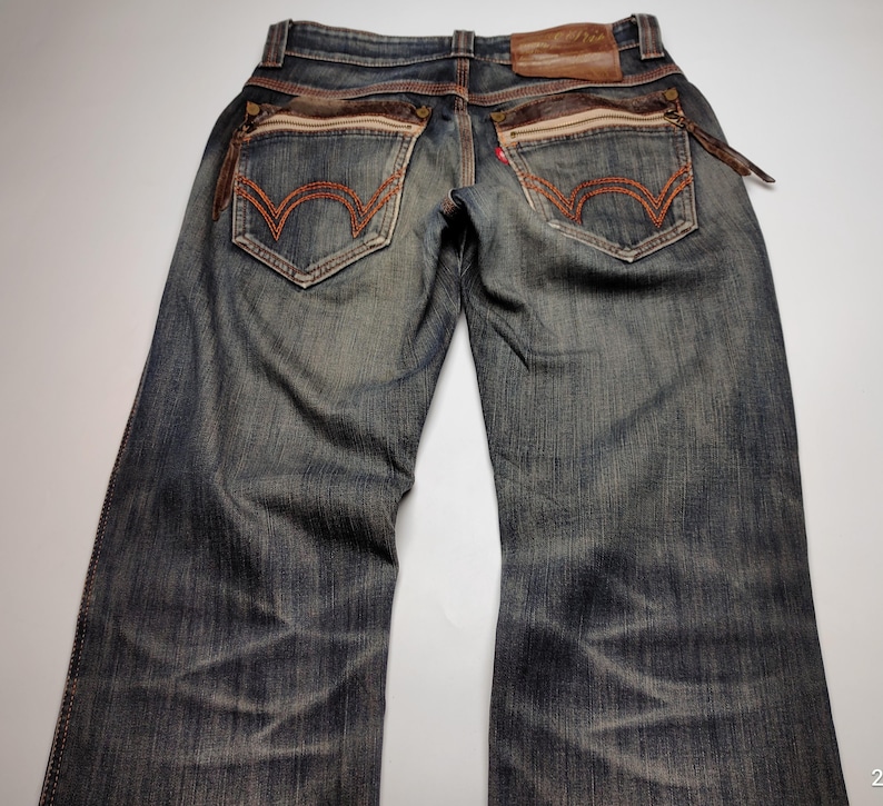 Puede incluir: Vaqueros de mezclilla de lavado oscuro con detalles en cuero marr&oacute;n y costuras. Incluyen bolsillos traseros decorativos con pespuntes naranjas, un parche de cuero marr&oacute;n y un detalle de cremallera. Los vaqueros tienen un aspecto descolorido y desgastado.