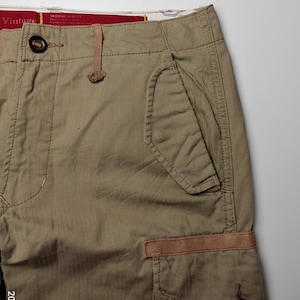 May include: Close-up of khaki-colored cargo pants. The pants feature multiple pockets, including a large side pocket with a button closure and a smaller pocket with a brown accent. The waistband has a button closure and a small loop. The word "Vintage" is visible on the waistband.