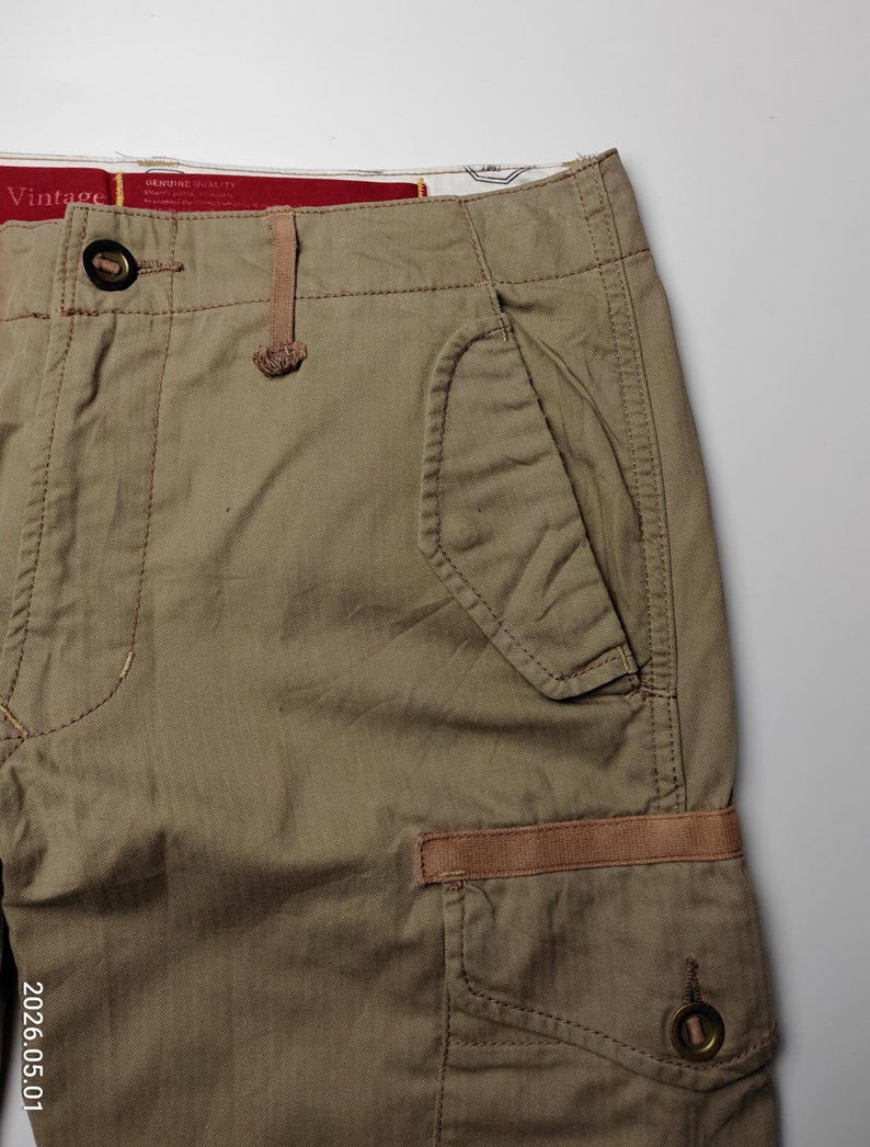 May include: Close-up of khaki-colored cargo pants. The pants feature multiple pockets, including a large side pocket with a button closure and a smaller pocket with a brown accent. The waistband has a button closure and a small loop. The word "Vintage" is visible on the waistband.