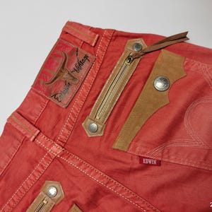 May include: Close-up of red denim jeans with decorative stitching and leather accents. Features include a zippered pocket, a leather patch with a bull graphic, and the brand name "Edwin" on a small red tag. The jeans have a vintage aesthetic.