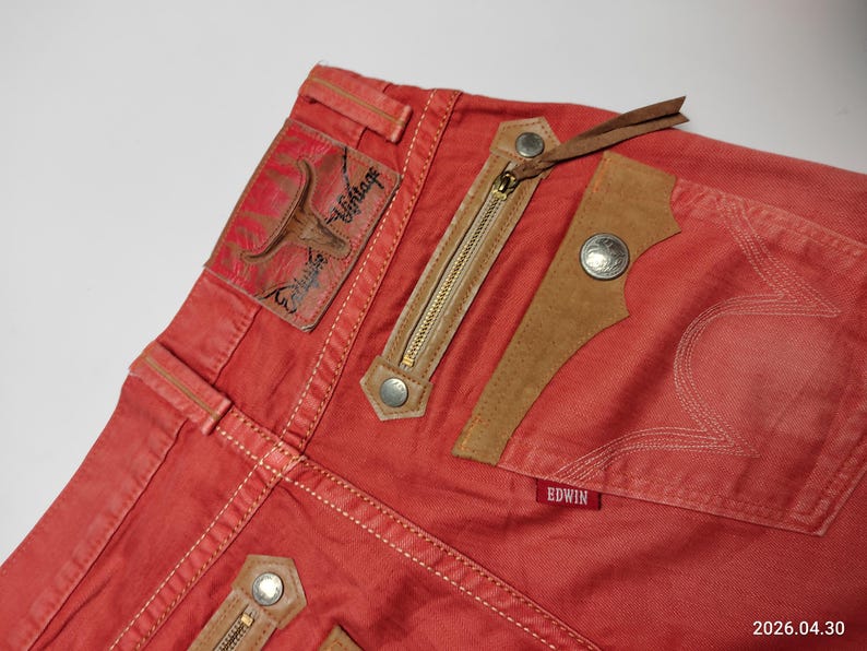 May include: Close-up of red denim jeans with decorative stitching and leather accents. Features include a zippered pocket, a leather patch with a bull graphic, and the brand name "Edwin" on a small red tag. The jeans have a vintage aesthetic.