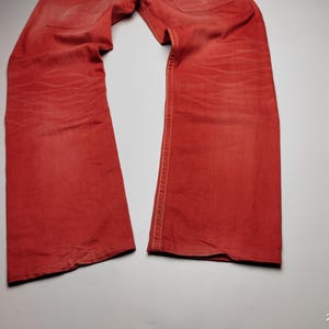 May include: A pair of red denim jeans, laid flat. The jeans have a classic five-pocket design and a straight leg cut. The fabric shows subtle fading and wear, adding character to the garment. The stitching is visible along the seams.