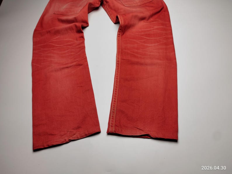 May include: A pair of red denim jeans, laid flat. The jeans have a classic five-pocket design and a straight leg cut. The fabric shows subtle fading and wear, adding character to the garment. The stitching is visible along the seams.