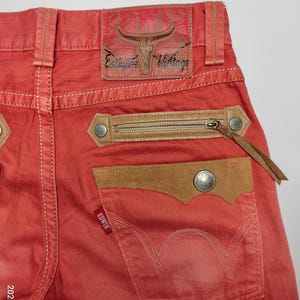May include: Close-up of red denim jeans with a brown leather patch and zipper pocket. The jeans feature a leather patch with a bull skull design and the words "Exclusive Vintage". The back pocket has the brand name "Edwin".
