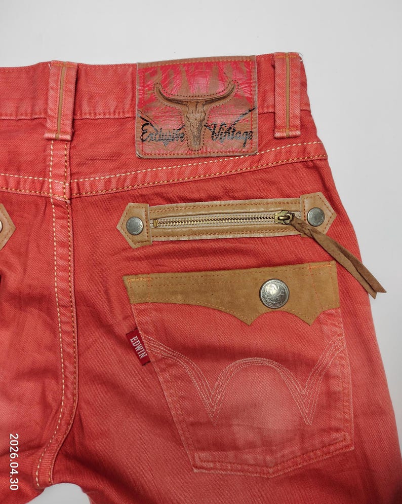 May include: Close-up of red denim jeans with a brown leather patch and zipper pocket. The jeans feature a leather patch with a bull skull design and the words "Exclusive Vintage". The back pocket has the brand name "Edwin".