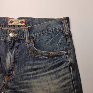 May include: Close-up of blue denim jeans with orange stitching. The jeans have a classic five-pocket design, a button closure, and a small decorative label on the back. The denim has a faded, worn look.