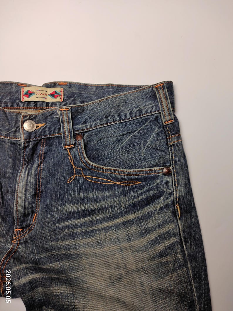 May include: Close-up of blue denim jeans with orange stitching. The jeans have a classic five-pocket design, a button closure, and a small decorative label on the back. The denim has a faded, worn look.