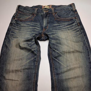 May include: Blue denim jeans with a faded wash and visible wear marks. The jeans have classic five-pocket styling, with copper-colored stitching and a button and zipper closure. The waistband features a small, decorative patch.