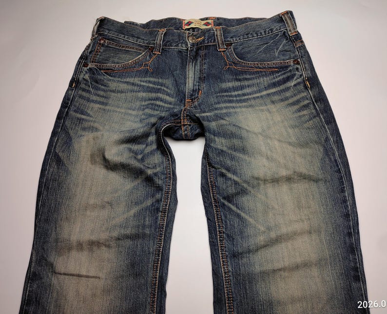 May include: Blue denim jeans with a faded wash and visible wear marks. The jeans have classic five-pocket styling, with copper-colored stitching and a button and zipper closure. The waistband features a small, decorative patch.