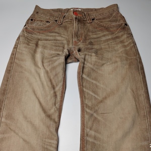 May include: Brown denim jeans with red stitching and a red button. The jeans have a faded, worn look with visible texture and pockets. The garment is laid flat, showcasing the details of the design.