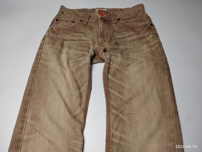 May include: Brown denim jeans with red stitching and a red button. The jeans have a faded, worn look with visible texture and pockets. The garment is laid flat, showcasing the details of the design.