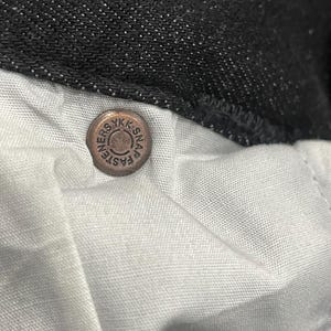 Puede incluir: Primer plano de un cierre de bot&oacute;n a presi&oacute;n de color cobre en unos vaqueros oscuros. El cierre de bot&oacute;n a presi&oacute;n circular tiene el texto "YKK SNAP FASTENERS". Los vaqueros tienen un forro de tela blanca.