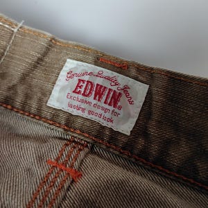 May include: Close-up of brown denim jeans with orange stitching. A white tag with red text reads "EDWIN Genuine Quality Jeans." The jeans have a textured appearance and are likely vintage or a classic style.