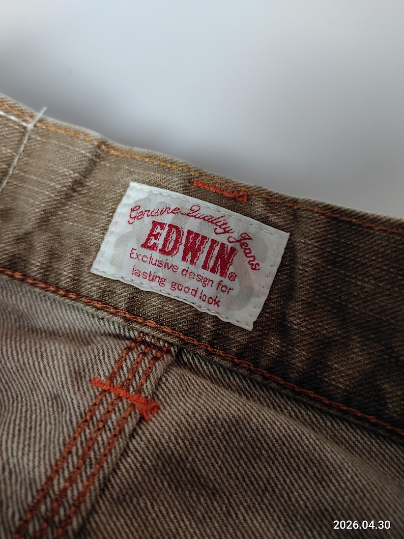 May include: Close-up of brown denim jeans with orange stitching. A white tag with red text reads "EDWIN Genuine Quality Jeans." The jeans have a textured appearance and are likely vintage or a classic style.