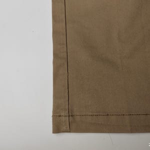 May include: Close-up of a section of tan fabric, showing a seam with dark thread. The fabric has a subtle texture and is folded over at the bottom, revealing a hem. The background is plain white.