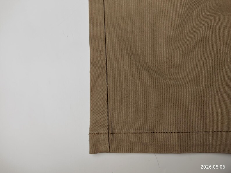 May include: Close-up of a section of tan fabric, showing a seam with dark thread. The fabric has a subtle texture and is folded over at the bottom, revealing a hem. The background is plain white.