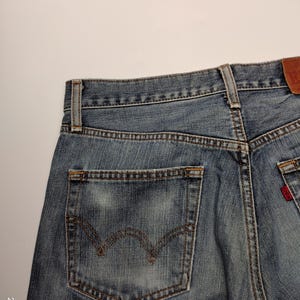 May include: Close-up of the back of blue denim jeans. The jeans have a classic five-pocket design, with a back pocket featuring a distinctive curved stitch. A small red tag is visible on the back pocket. The jeans are faded and worn.