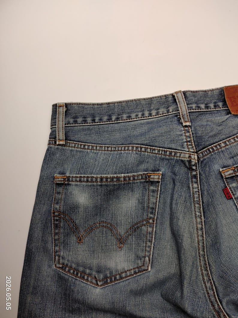 May include: Close-up of the back of blue denim jeans. The jeans have a classic five-pocket design, with a back pocket featuring a distinctive curved stitch. A small red tag is visible on the back pocket. The jeans are faded and worn.