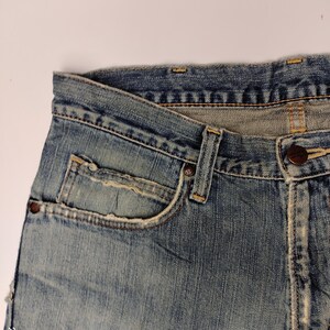 May include: Close-up of a pair of light blue denim jeans. The jeans have a classic five-pocket design, with a small pocket on the front and two pockets on the back. The denim has a faded, worn look, with visible stitching and a button closure.