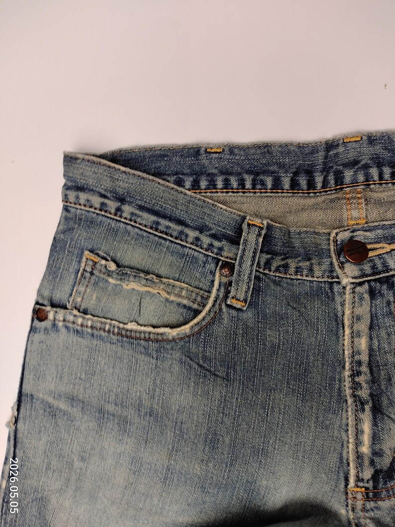 May include: Close-up of a pair of light blue denim jeans. The jeans have a classic five-pocket design, with a small pocket on the front and two pockets on the back. The denim has a faded, worn look, with visible stitching and a button closure.