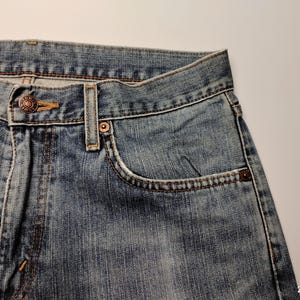 May include: Close-up of blue denim jeans. The jeans feature a button closure, a small pocket, and visible stitching. The denim has a faded appearance, with a worn look.