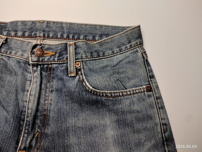 May include: Close-up of blue denim jeans. The jeans feature a button closure, a small pocket, and visible stitching. The denim has a faded appearance, with a worn look.