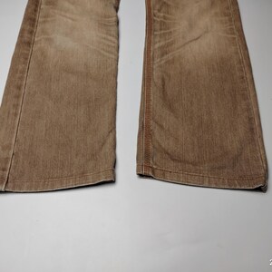 May include: Close-up of the lower portion of a pair of brown denim jeans. The jeans have a faded appearance and visible stitching along the seams. The fabric appears to be a medium weight denim.